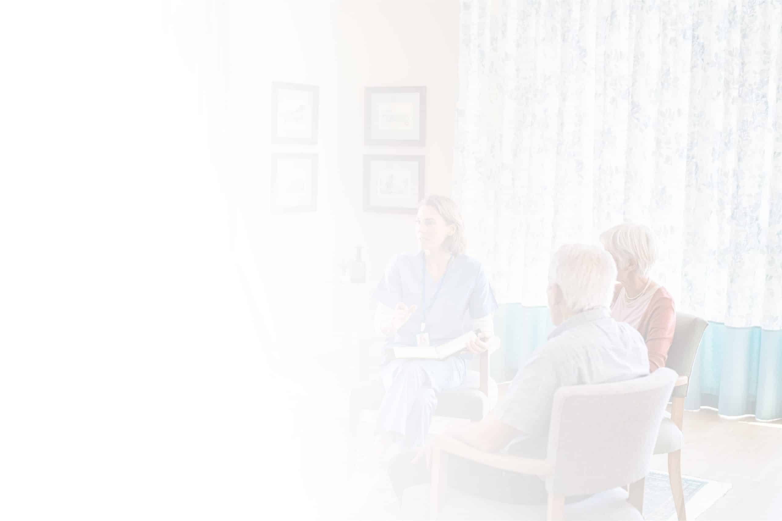 A healthcare professional in blue scrubs speaks with an elderly couple seated in a bright, airy room with light curtains and framed pictures on the wall. The image, softened by a vignette effect, captures a heartwarming moment at North Pines Assisted Living.