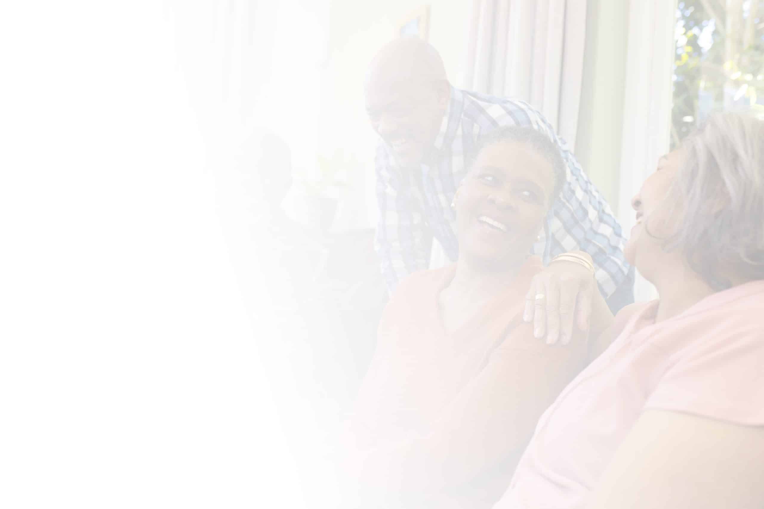 A light, hazy image of three people smiling and interacting indoors at North Pines Assisted Living. Two women are seated, facing each other and smiling, while a standing man leans forward with a hand on one woman's shoulder. Bright natural light streams in from the window behind them.