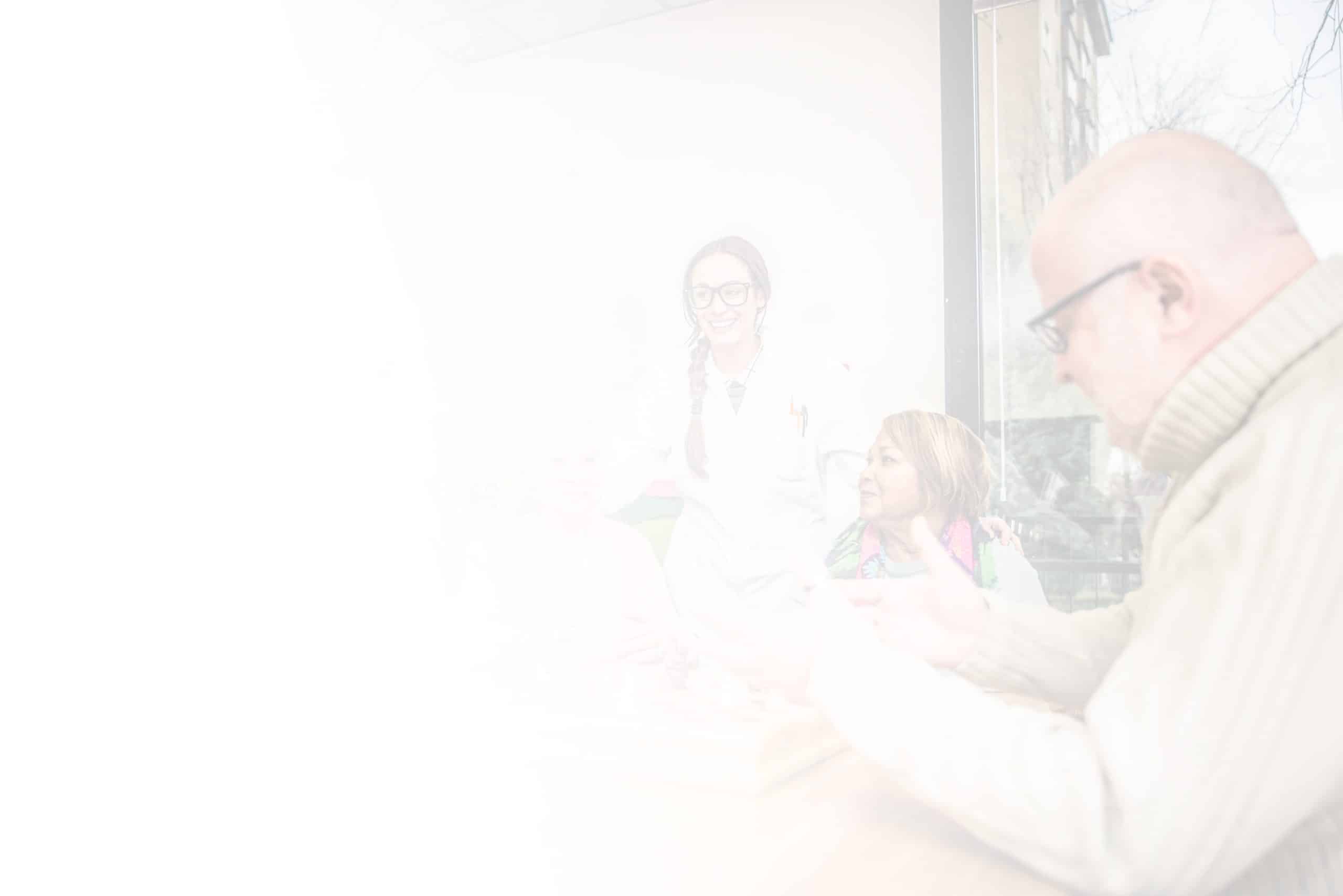 A blurred image features an elderly man and woman sitting at a table, possibly playing a game, in North Pines Assisted Living. A female medical professional is standing beside them, smiling. Another person is partially visible at the edge of the image. They appear to be in a well-lit room.