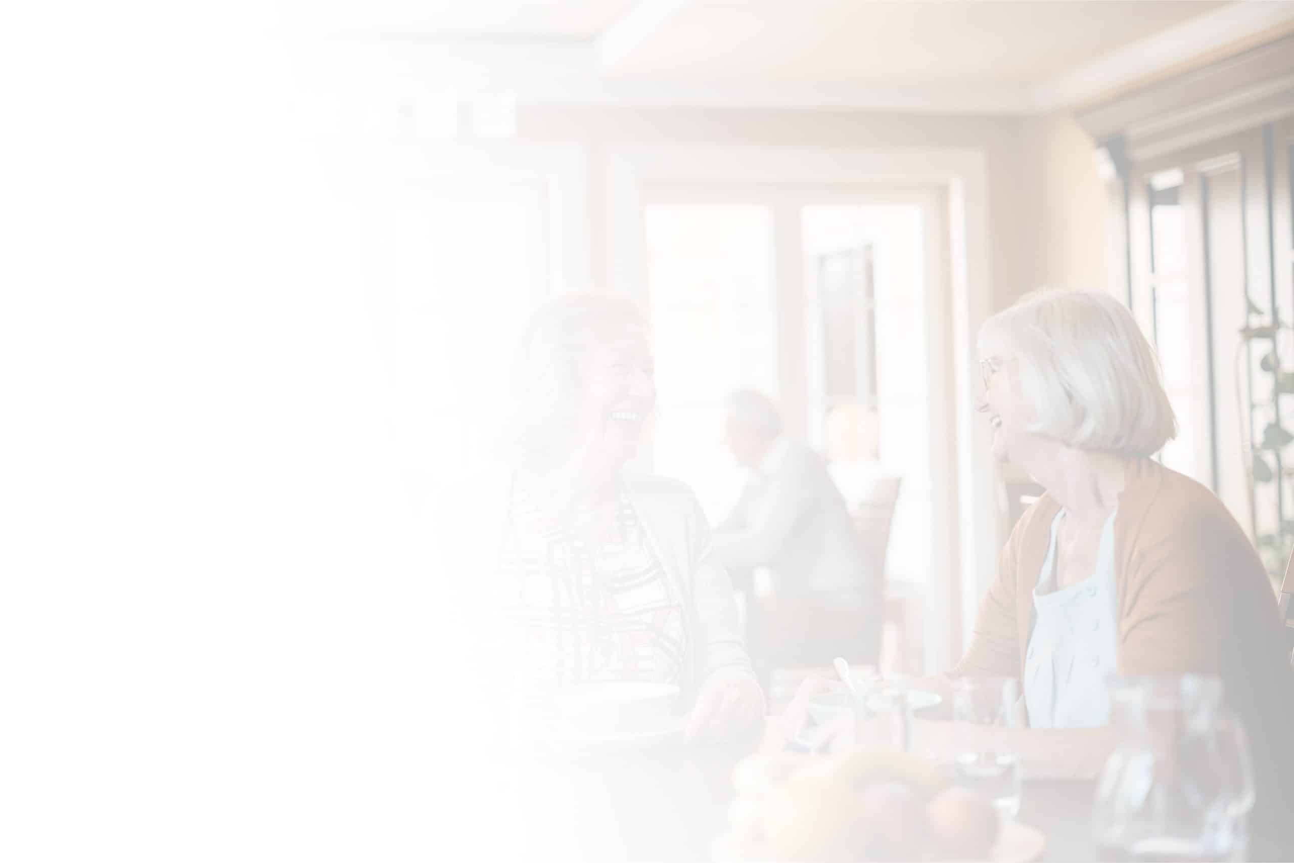 Two older women are sitting at a table, engaging in a lively conversation and laughing. The background shows a bright room with large windows, filled with natural light, typical of North Pines Assisted Living. The image is partially faded, giving it a soft, ethereal feel.