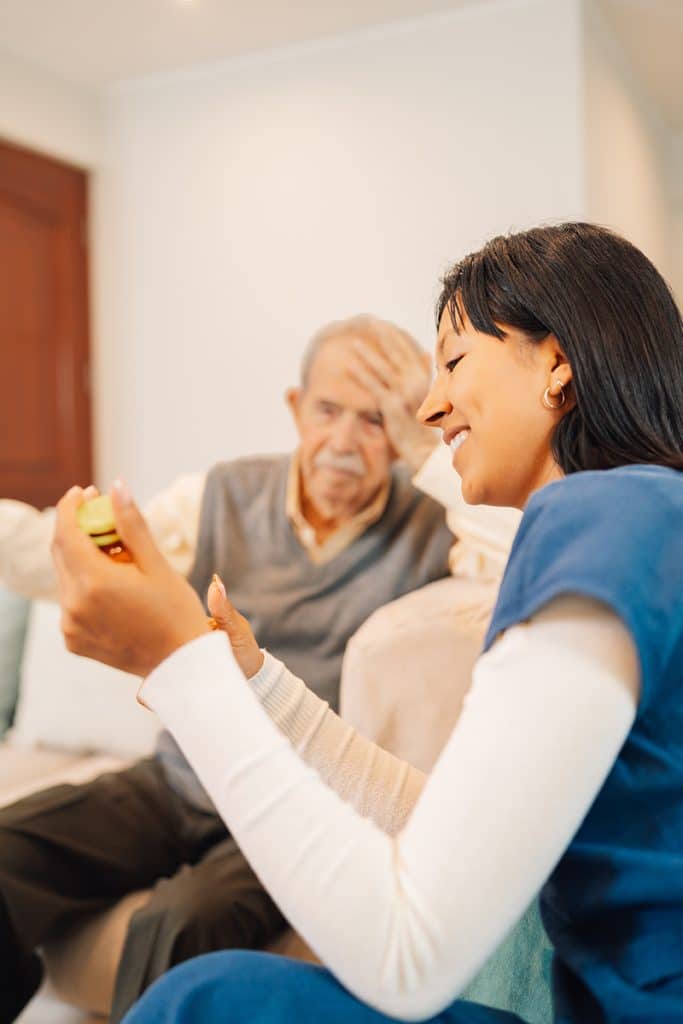 A woman, smiling and wearing a blue top, sits next to an elderly man who is touching his forehead and looking concerned. She holds a small, yellow container in her hand. They are indoors at North Pines Assisted Living, sitting on a couch in a well-lit room.