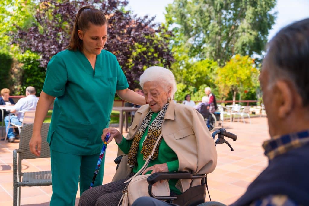 A caregiver in a green uniform assists an elderly woman in a wheelchair outside at North Pines Assisted Living. The elderly woman, holding a cane and wearing a scarf, smiles as she enjoys the fresh air. Other residents are visible in the background, enjoying the trees and greenery of the senior living facility.