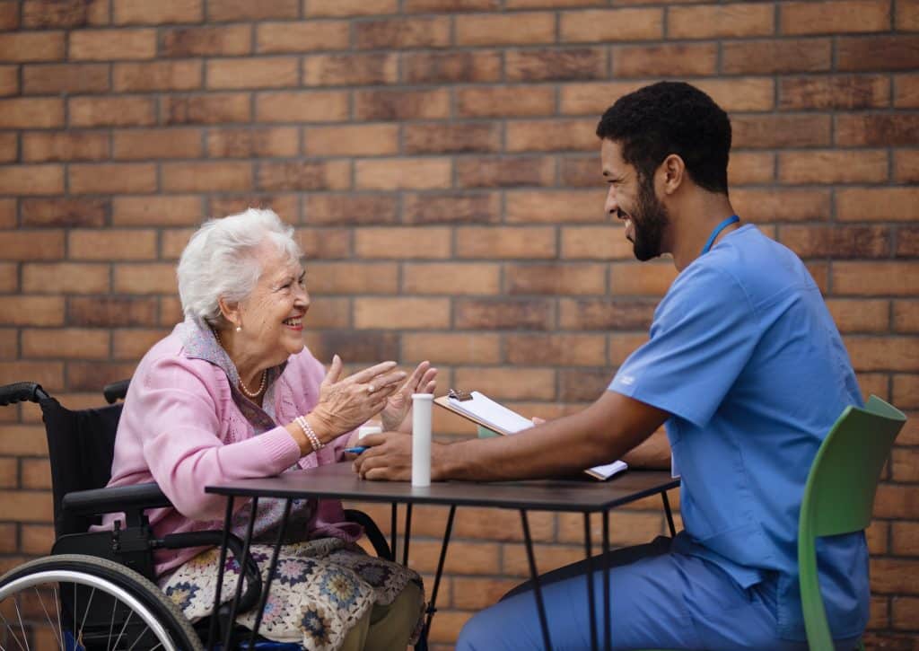 An elderly woman in a wheelchair, wearing a pink cardigan, sits at a table with a male nurse in blue scrubs at North Pines Assisted Living. They are both smiling and appear to be having a cheerful conversation. A clipboard and a water bottle are on the table. They are outdoors.