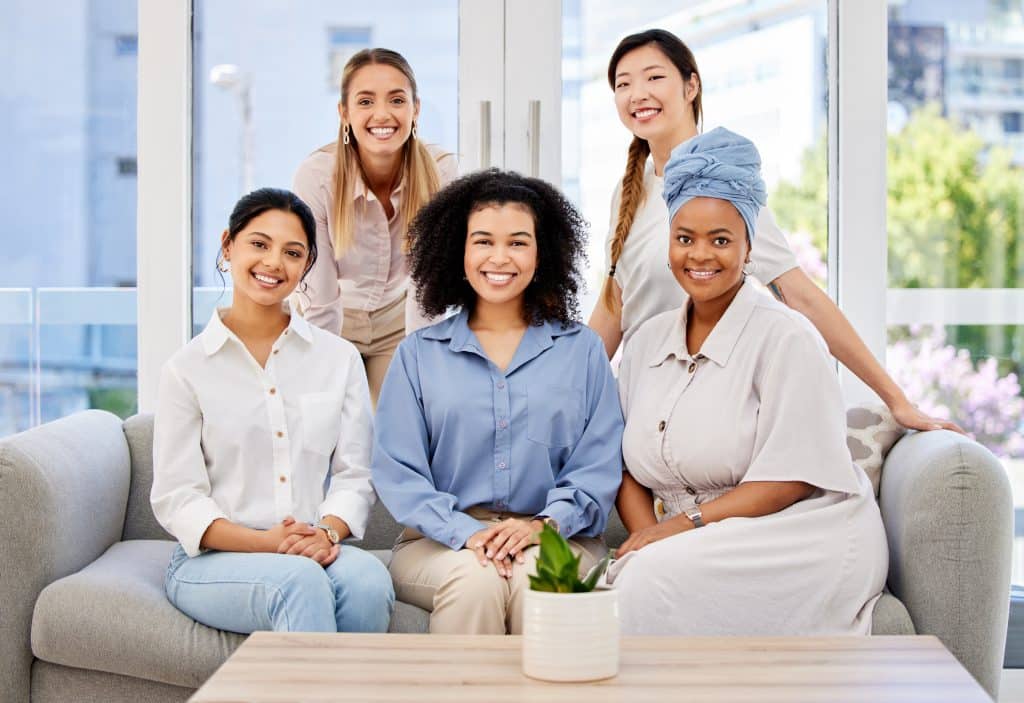 Five women are smiling and sitting together on a light-colored couch in a well-lit room at North Pines Assisted Living. They appear happy and relaxed, wearing casual and semi-casual outfits. In front of them is a wooden coffee table with a small potted plant.