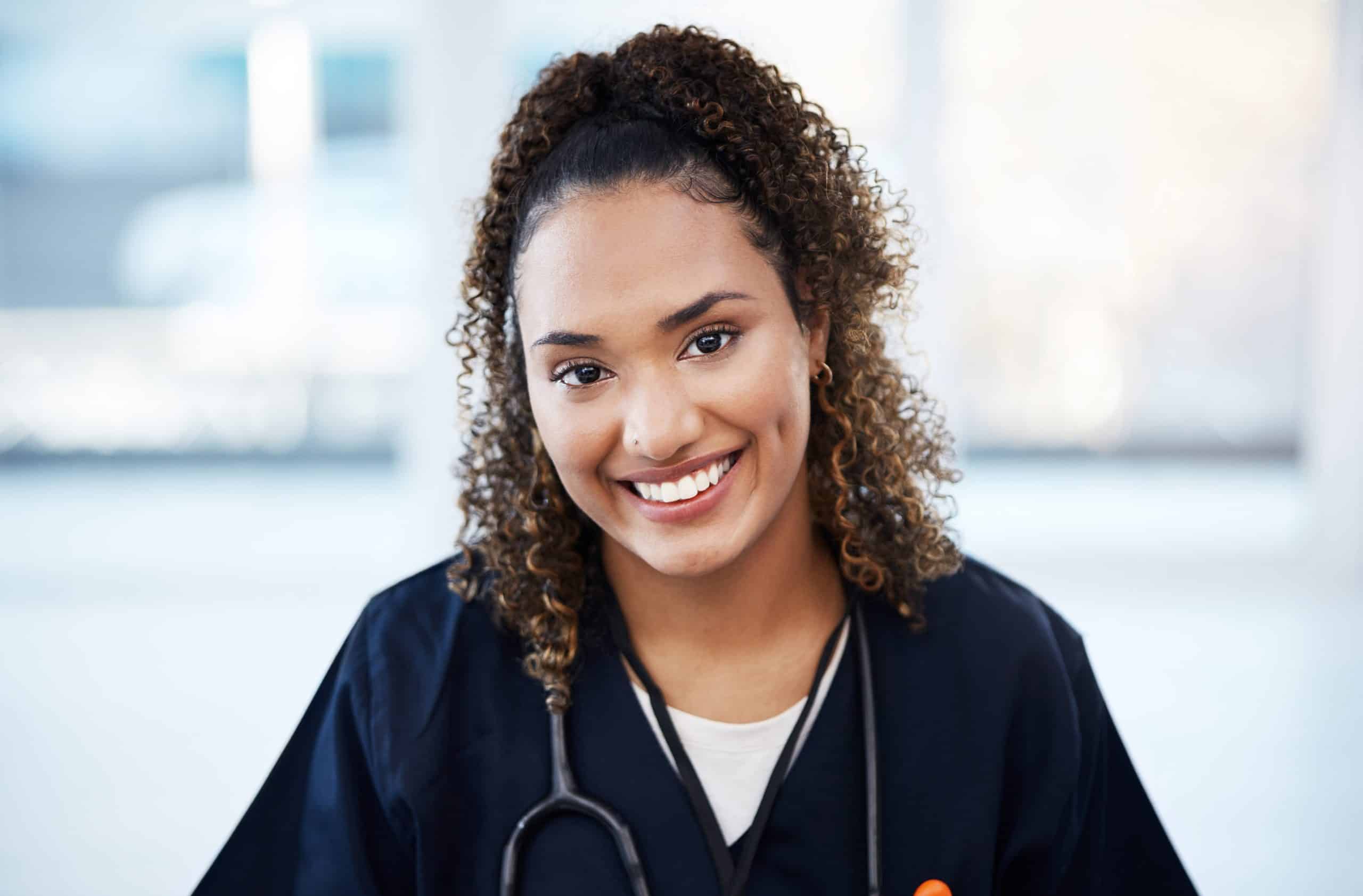 A smiling healthcare worker wearing navy blue scrubs, a white undershirt, and a stethoscope around her neck stands in a bright setting at North Pines Assisted Living. She has curly hair pulled back and is looking directly at the camera. The background is softly blurred.