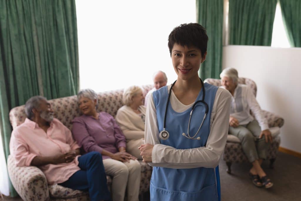 A healthcare worker wearing scrubs and a stethoscope stands smiling with arms crossed in the foreground. In the background, a group of elderly individuals are seated on couches, engaging in conversation and laughter. The room at North Pines Assisted Living has green curtains and a bright atmosphere.