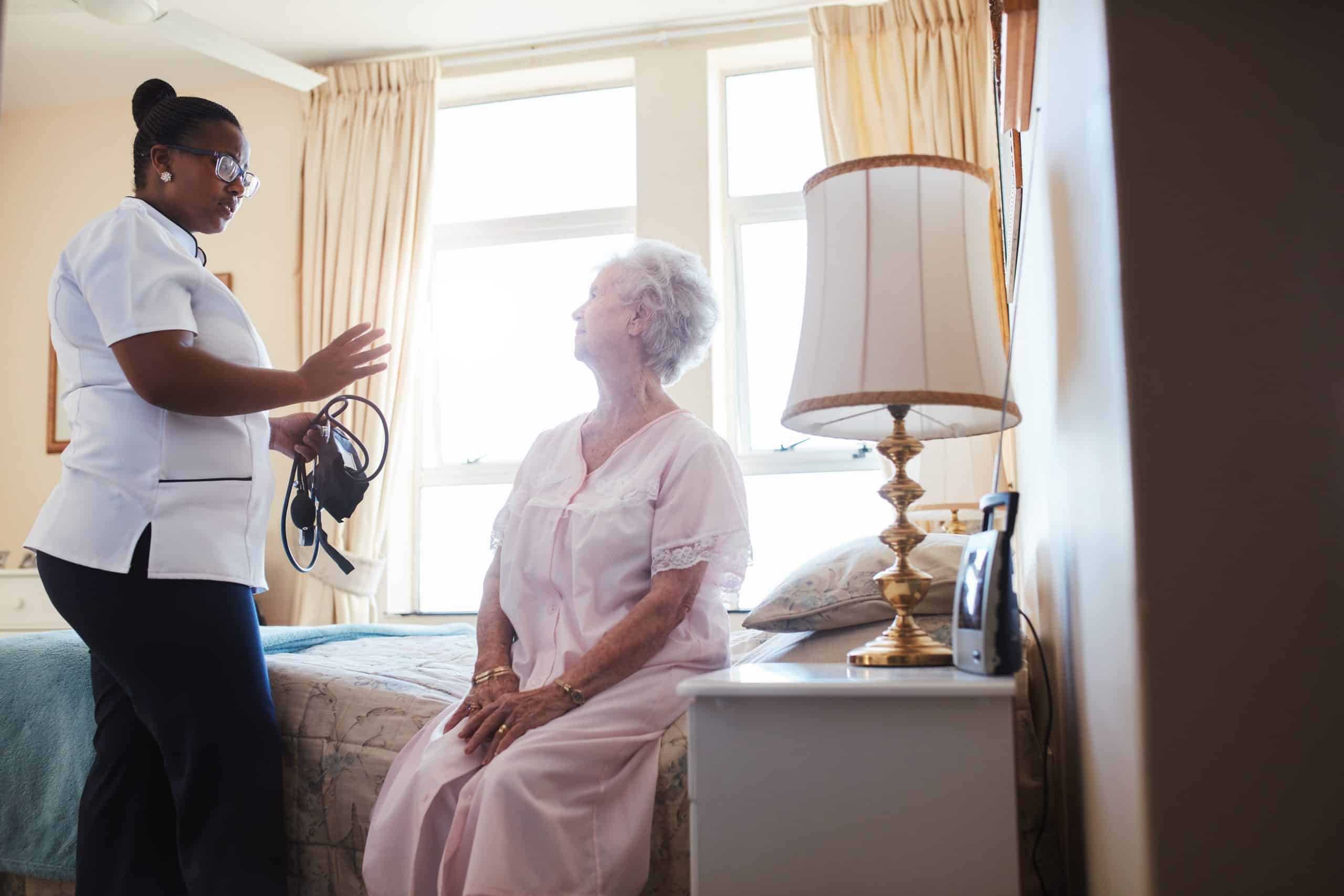 A nurse in a white uniform stands and gestures while speaking to an elderly woman sitting on a bed in a well-lit room at North Pines Assisted Living. The woman is wearing a light pink nightgown. The room has beige walls, a bedside table with a lamp, and a window with cream-colored curtains.