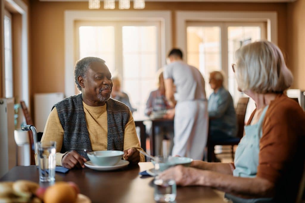 Elderly man and woman sitting at a dining table, enjoying a meal together in a warmly lit room of North Pines Assisted Living; other seniors and a caregiver can be seen in the background. Bright windows let in natural light, creating a cozy atmosphere.