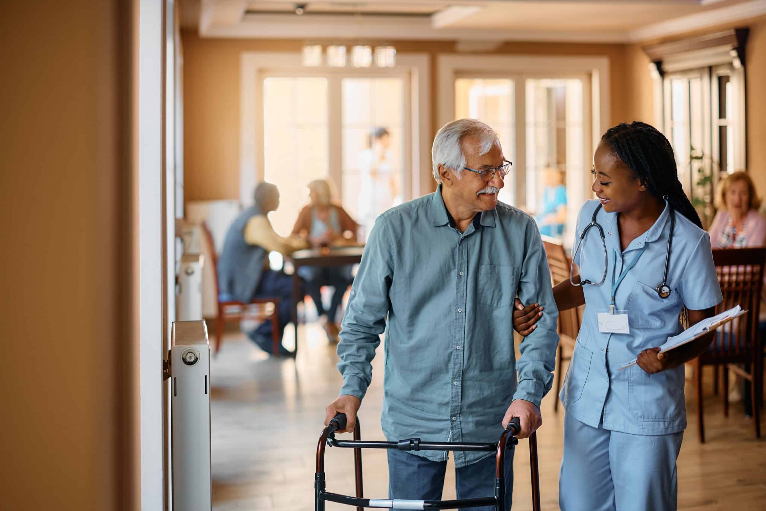 A smiling nurse assists an elderly man using a walker through the warmly lit halls of North Pines Assisted Living. In the background, other residents and staff are engaged in activities and socializing at tables, creating a cozy and supportive atmosphere at this senior living facility.