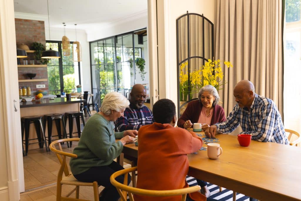 Five older adults sit around a dining table, engaged in an activity together. A bright, modern kitchen is visible in the background at North Pines Assisted Living. The setting is well-lit with large windows, and yellow flowers are arranged on the table, creating a warm and welcoming atmosphere.