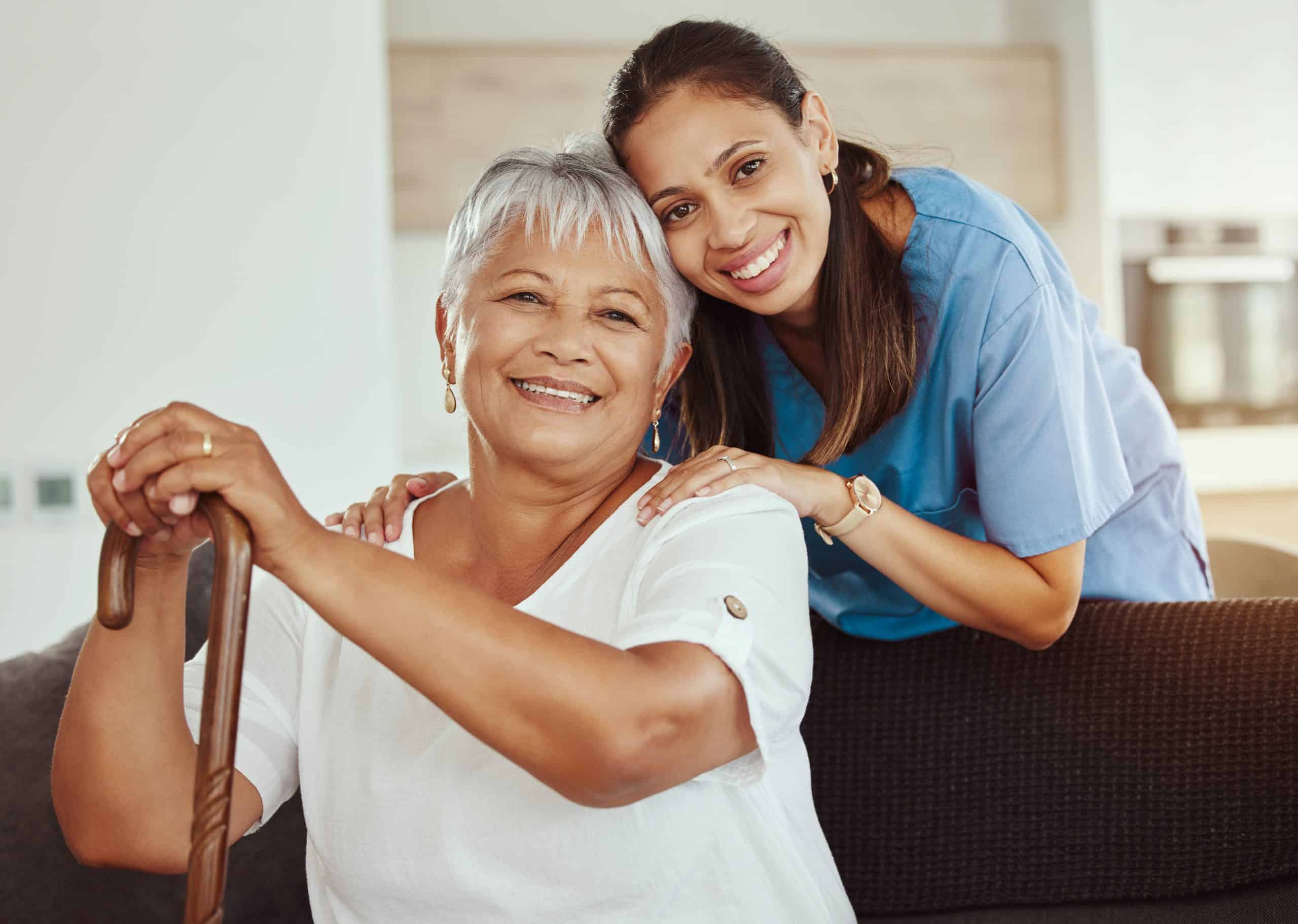 An elderly woman with short gray hair sits on a couch, smiling and holding a cane. A younger woman, wearing blue scrubs, stands behind her with hands gently placed on the elderly woman's shoulders, also smiling. They appear to be in a bright, cozy room at North Pines Assisted Living.