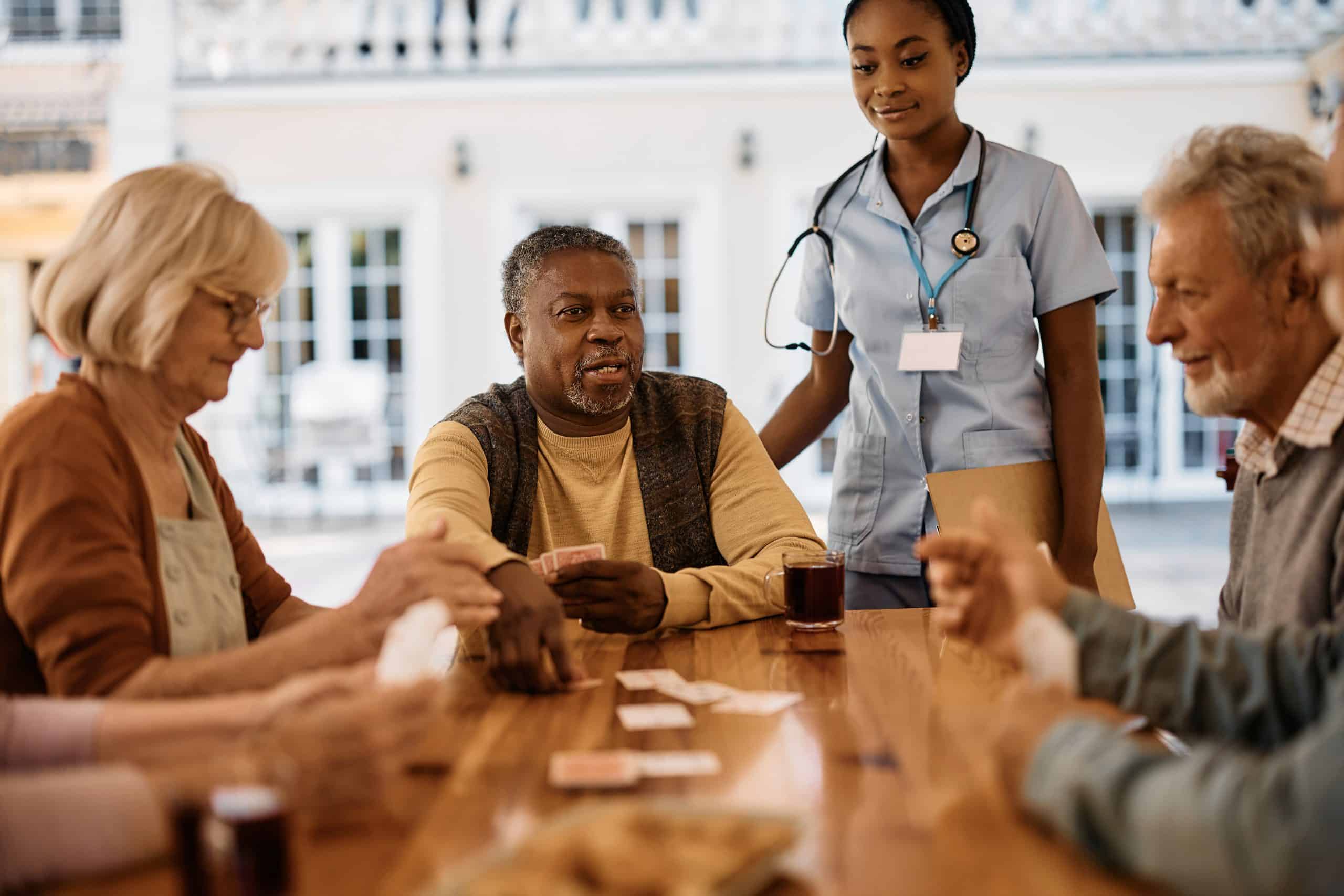 A group of senior adults is gathered around a wooden table playing cards on the outdoor patio of North Pines Assisted Living. A healthcare worker stands behind them, dressed in a blue uniform and holding a clipboard. Everyone is engaged and appears to be enjoying the activity.