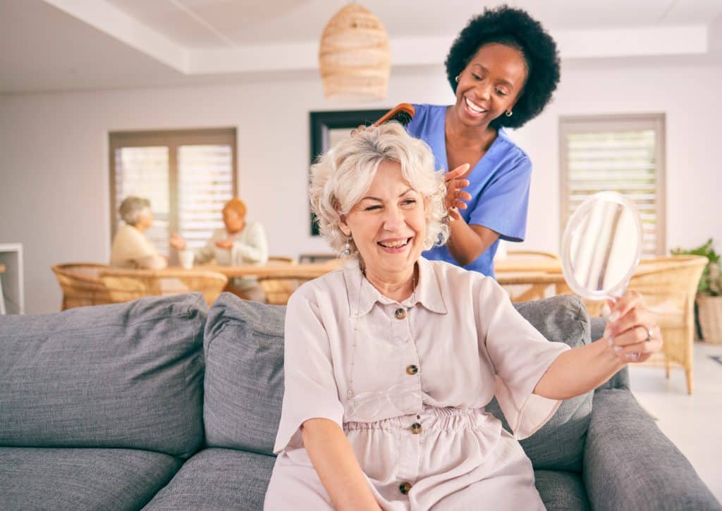 A smiling elderly woman sits on a couch at North Pines Assisted Living, holding a mirror as a caregiver combs her hair. In the background, two other residents are seated at a table with drinks. The setting appears to be a cozy, well-lit living area.