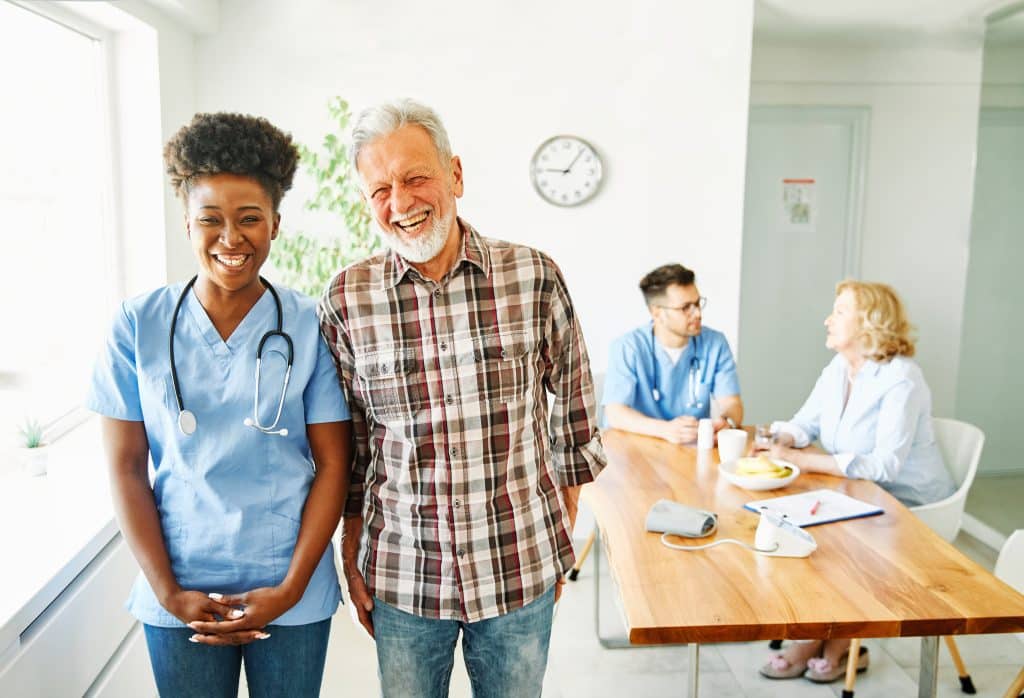 A smiling nurse with a stethoscope stands next to an elderly man wearing a plaid shirt in a bright room at North Pines Assisted Living. In the background, two other healthcare workers, a man and a woman, sit at a table and engage in conversation. A clock hangs on the wall behind them.