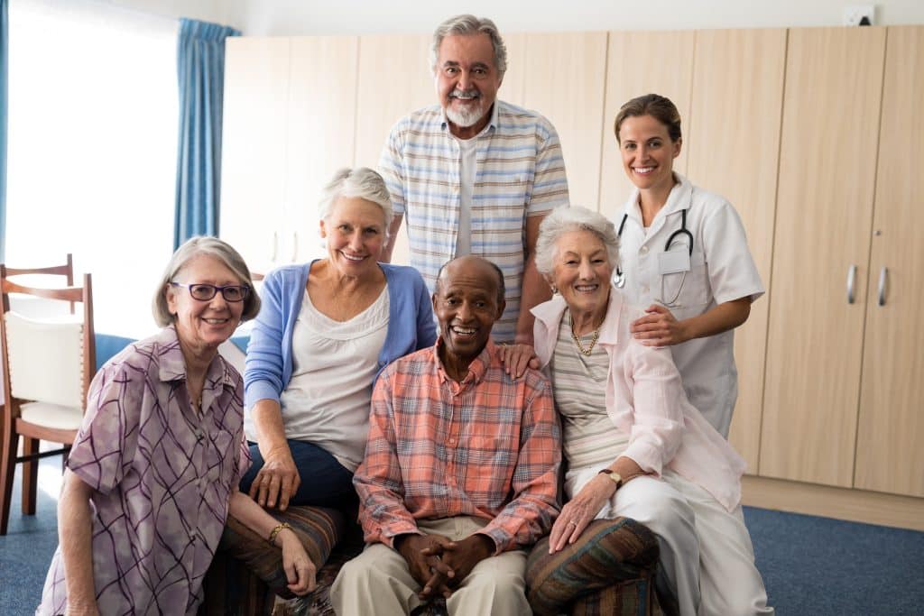 At North Pines Assisted Living, a group of elderly people is seated and smiling, accompanied by a male staff member standing behind them. A female medical professional in a white coat is standing to the right. The background shows a room with beige cabinets and a window with blue curtains.