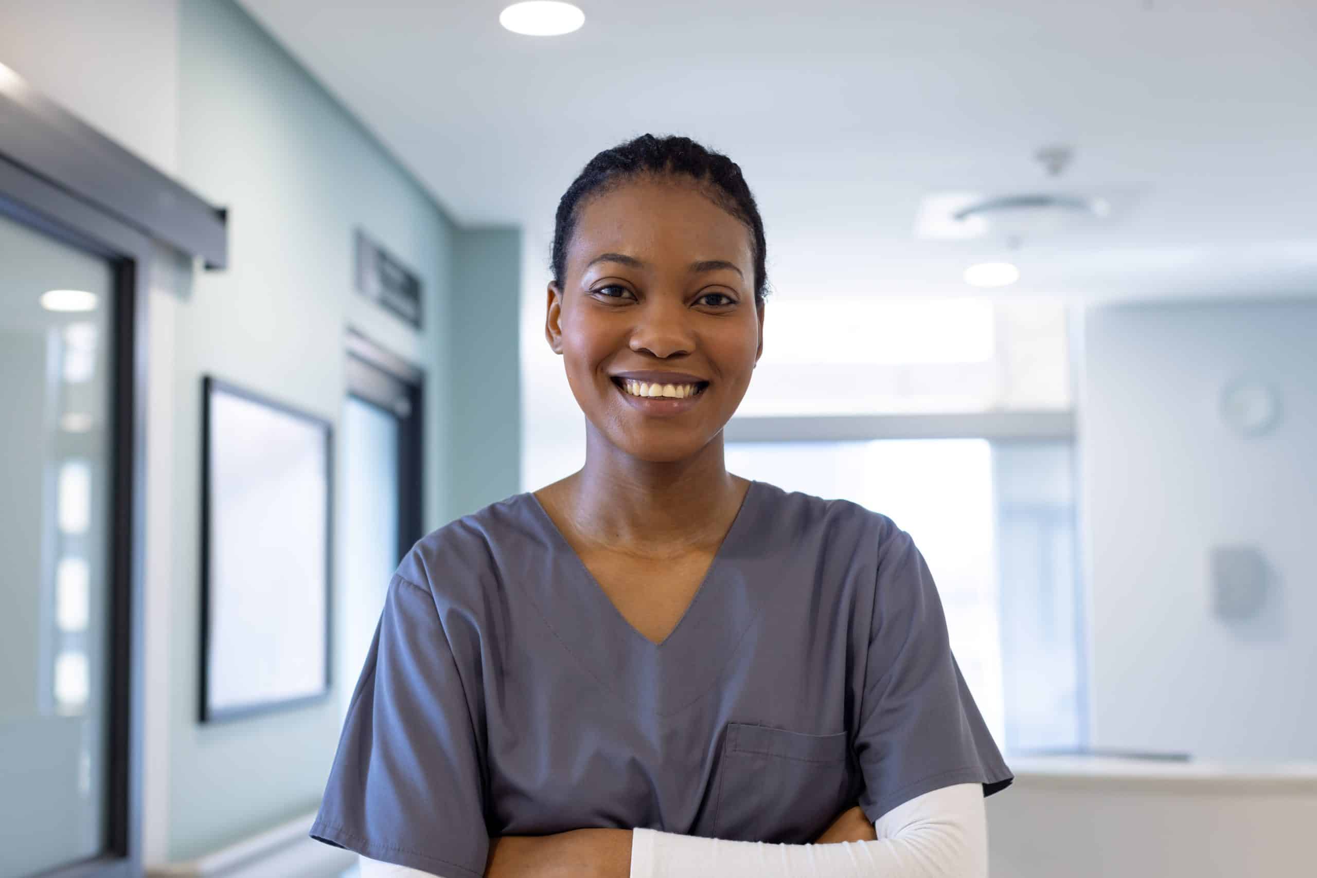 A smiling healthcare professional wearing gray scrubs stands with crossed arms in the bright corridor of North Pines Assisted Living.