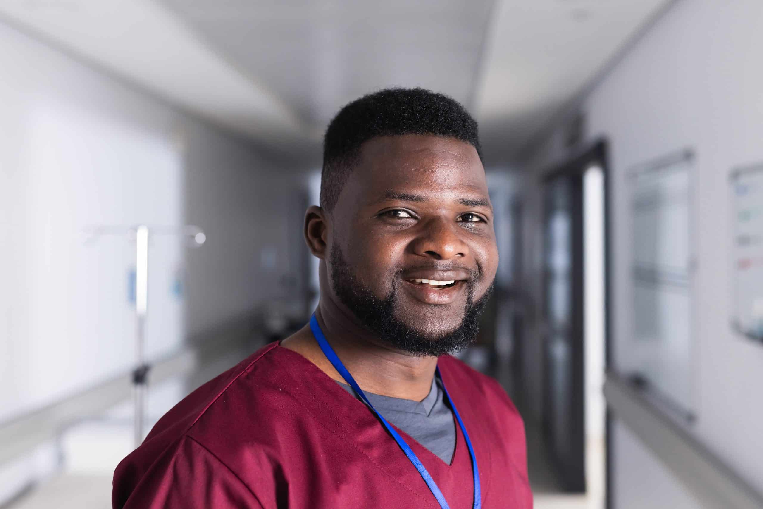 A smiling man wearing scrubs with a lanyard stands in a brightly lit hallway, possibly at North Pines Assisted Living, showcasing the welcoming environment of this senior living facility.