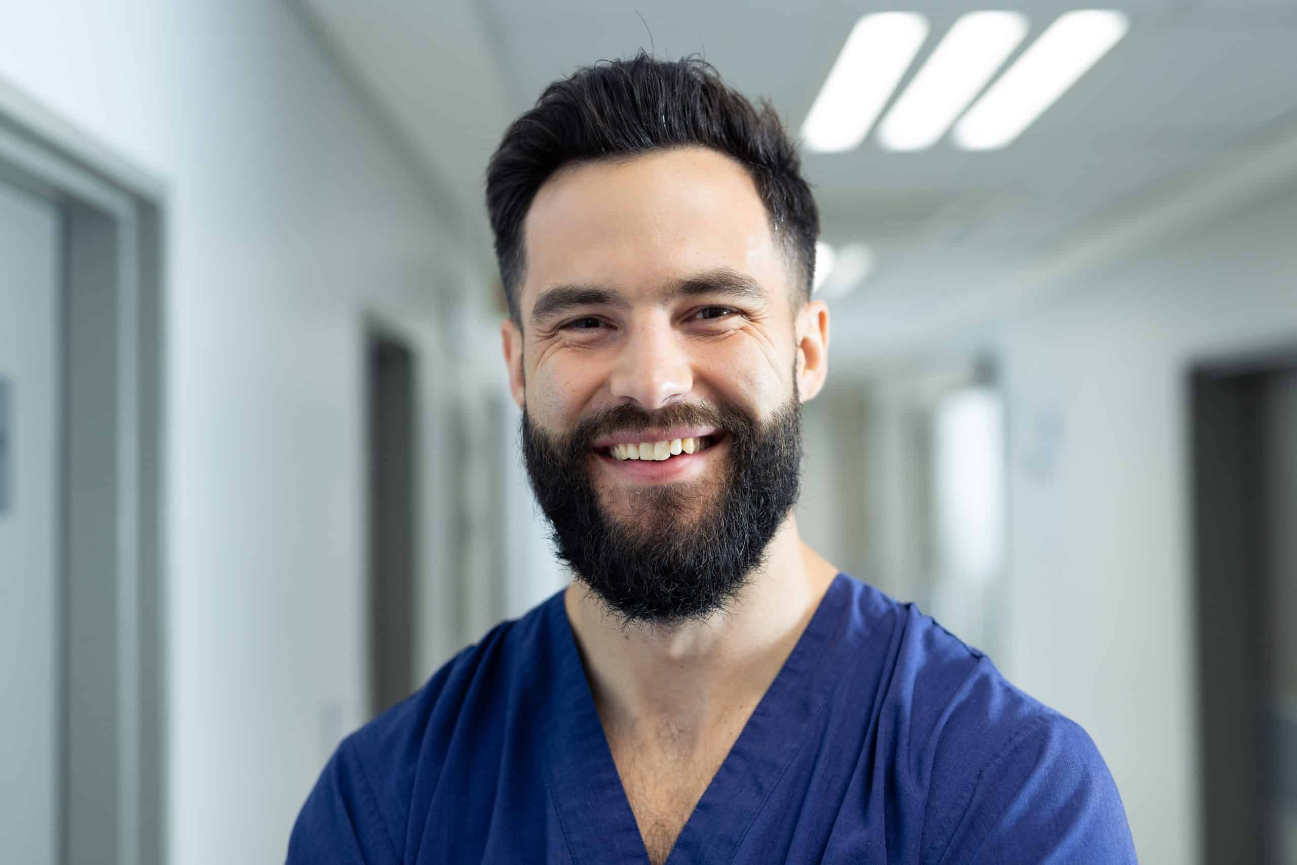 A bearded man wearing navy blue scrubs stands in a bright, modern hallway of North Pines Assisted Living, smiling warmly at the camera. The background features blurred doorways and overhead fluorescent lights, suggesting a welcoming and professional senior living facility.