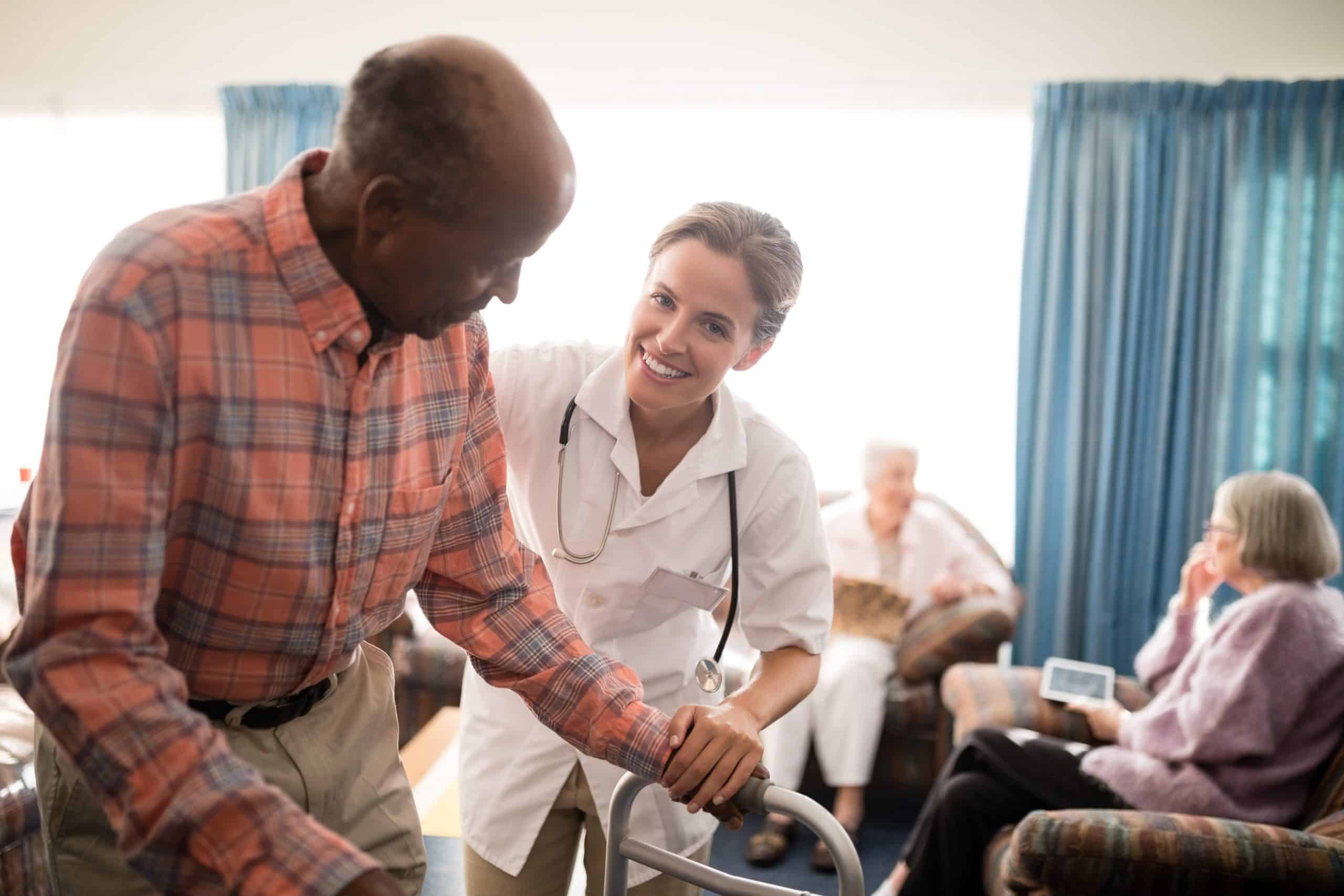 A healthcare worker assists an elderly man using a walker in a brightly lit room with blue curtains at North Pines Assisted Living. In the background, other elderly individuals are sitting on chairs, engaging in various activities. The healthcare worker is smiling and wearing a stethoscope.