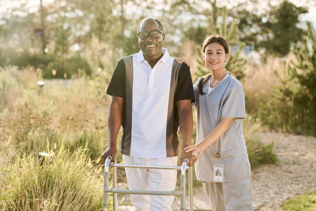 A smiling man with glasses and a black-and-white shirt uses a walker while standing next to a woman in scrubs, who is also smiling and has her hand on his shoulder. They are outdoors at North Pines Assisted Living on a sunny day, surrounded by greenery.