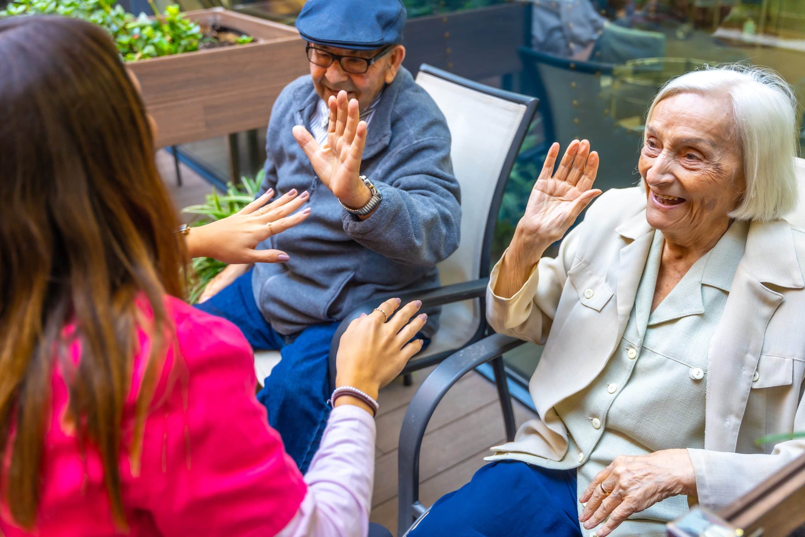 A young woman interacts with two elderly people, a man and a woman, seated on a patio chair at North Pines Assisted Living. Both seniors are smiling and raising their hands, appearing to engage in a playful activity. The scene is lively with greenery in the background.