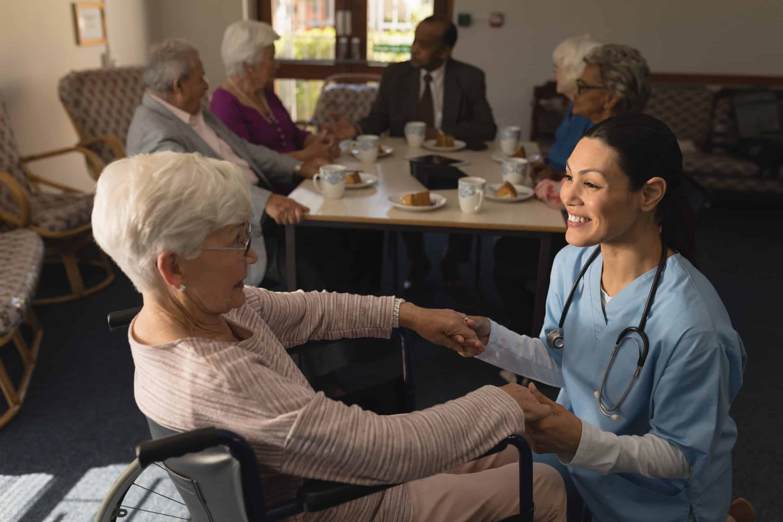 A nurse in blue scrubs and a stethoscope holds hands with a smiling elderly woman in a wheelchair at North Pines Assisted Living. In the background, a group of seniors sits around a table, engaging in conversation and enjoying drinks and snacks.